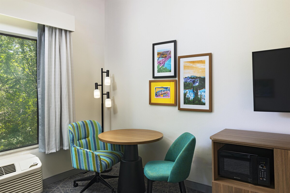 A room with a window, a wall-mounted flat-screen TV, a small round table, two chairs, and a microwave.
