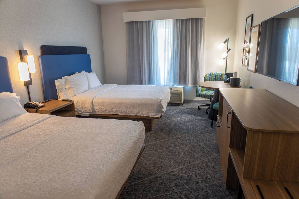 A hotel room with two beds, a desk, a chair, a lamp, and a window with curtains.