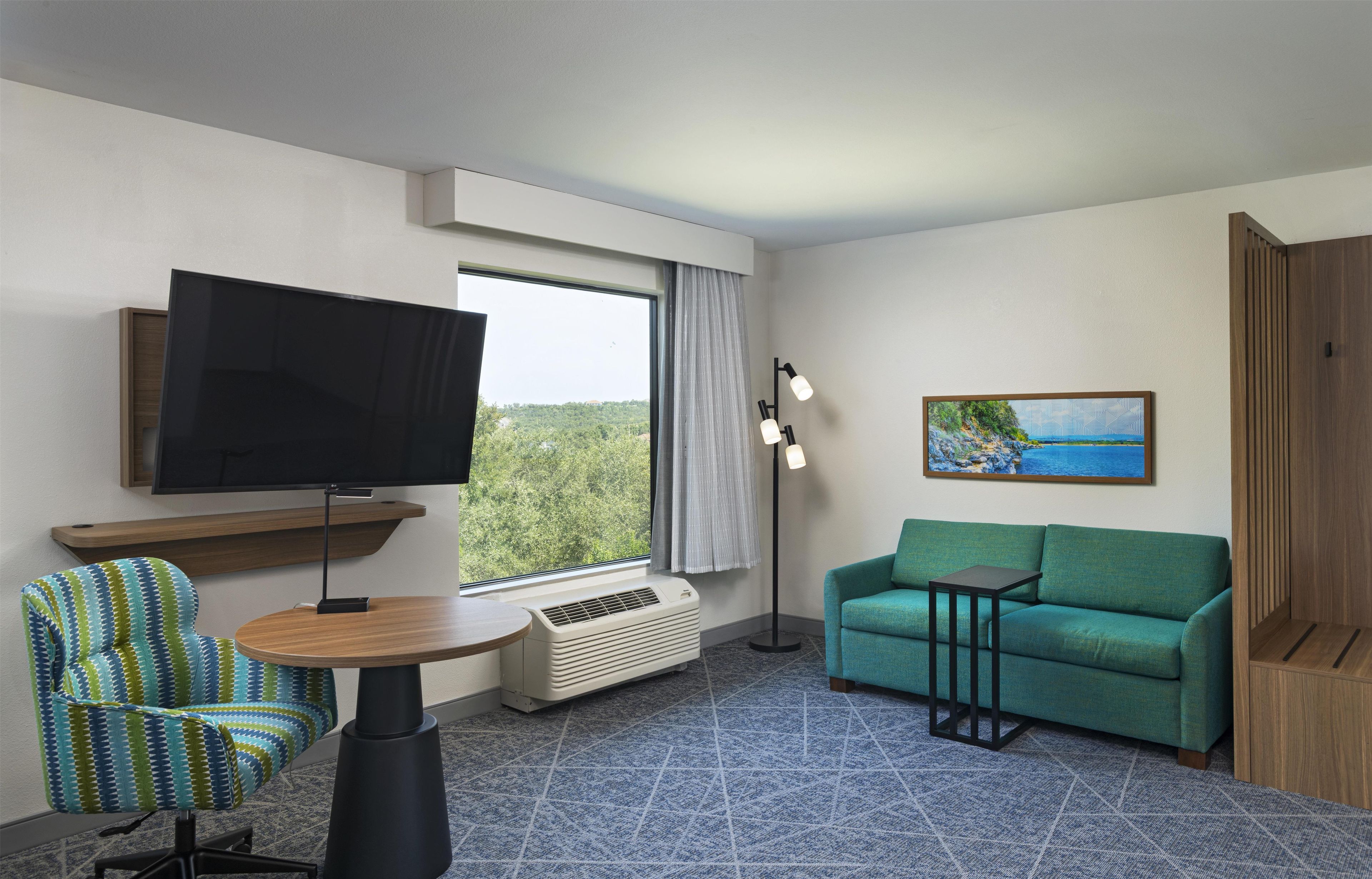 A hotel room with a television, a sofa, a chair, a small table, a floor lamp, and a painting on the wall.