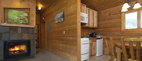 Interior de una cabaña de troncos con chimenea, paredes de madera, una zona de cocina y una mesa de comedor con sillas.