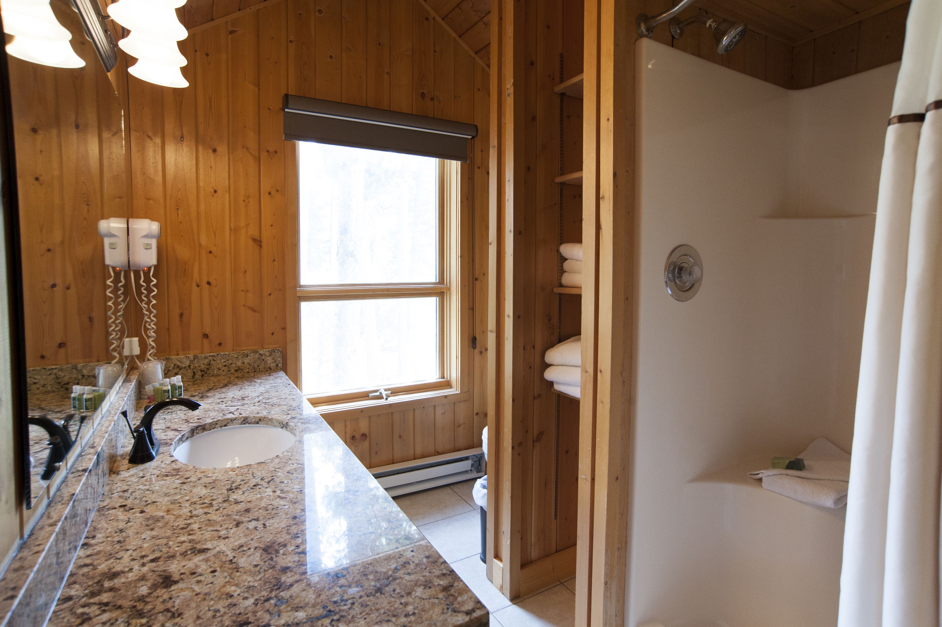 A bathroom with a granite countertop, a double sink, a mirror, a window, a shower, and wooden walls.