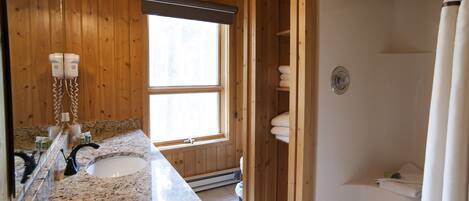 A bathroom with a granite countertop, a double sink, a mirror, a window, a shower, and wooden walls.