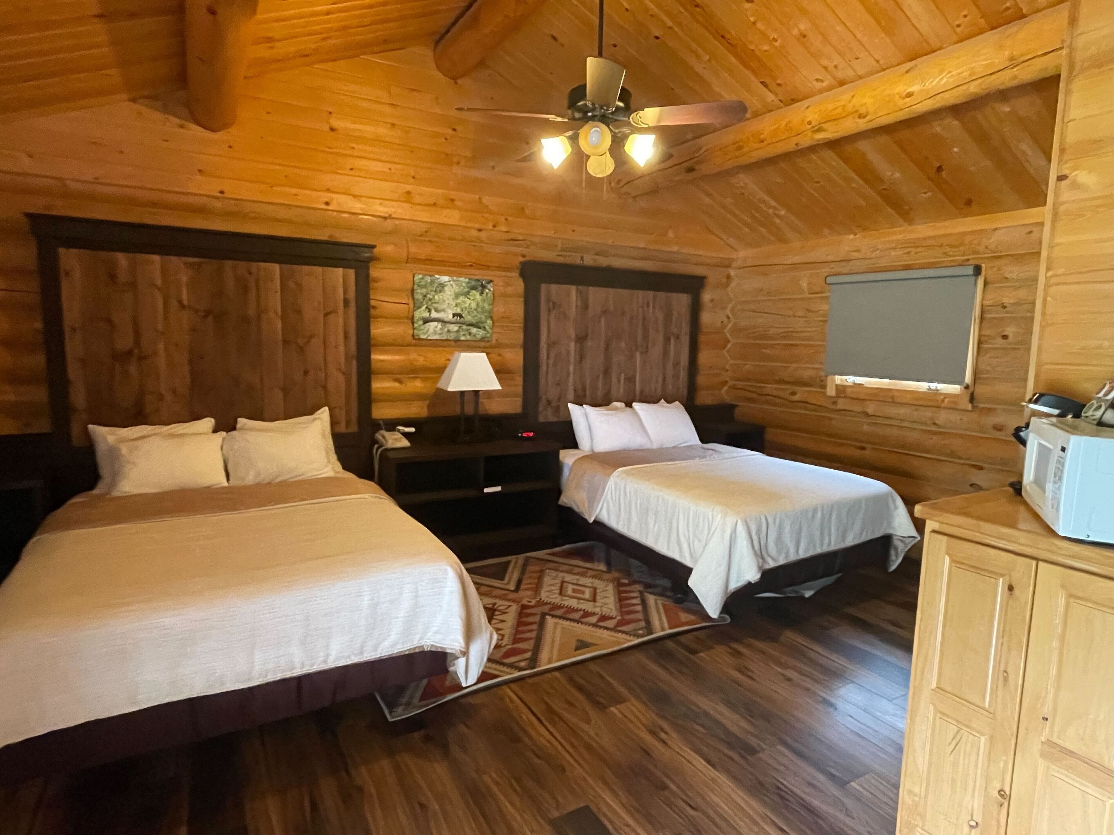 A wooden cabin bedroom with two beds, a ceiling fan, a nightstand, a lamp, and a microwave.