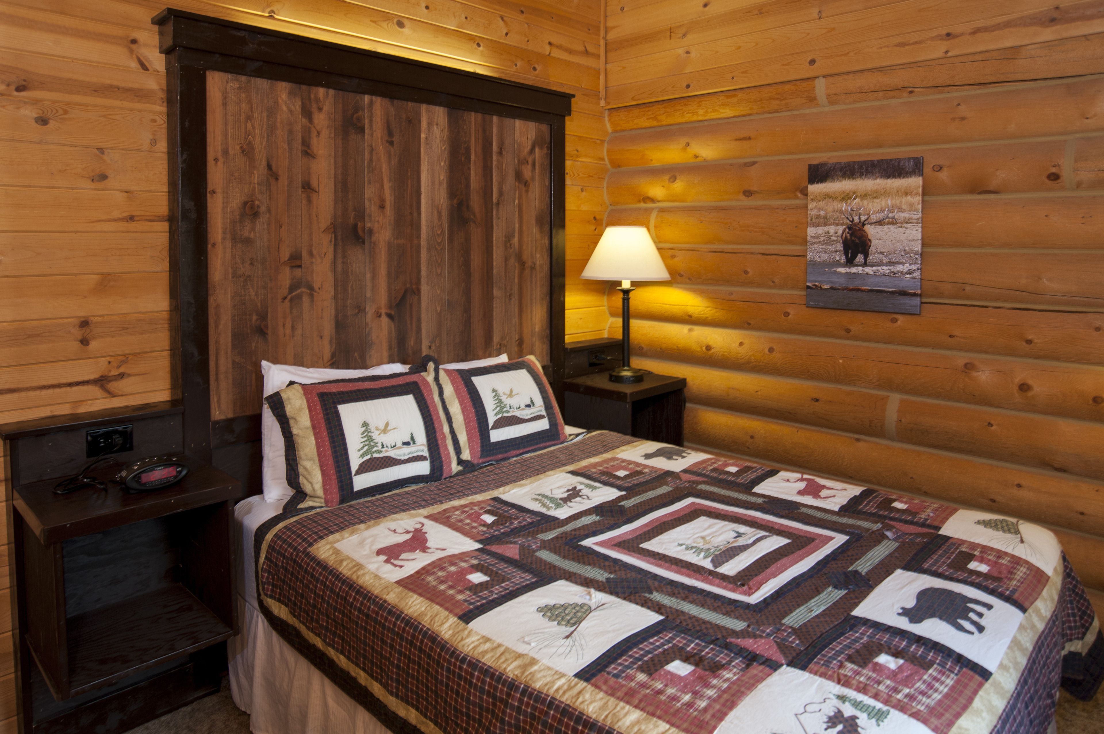 A log cabin room with a wooden headboard, a quilted bedspread, a nightstand with a lamp, and a framed picture on the wall.