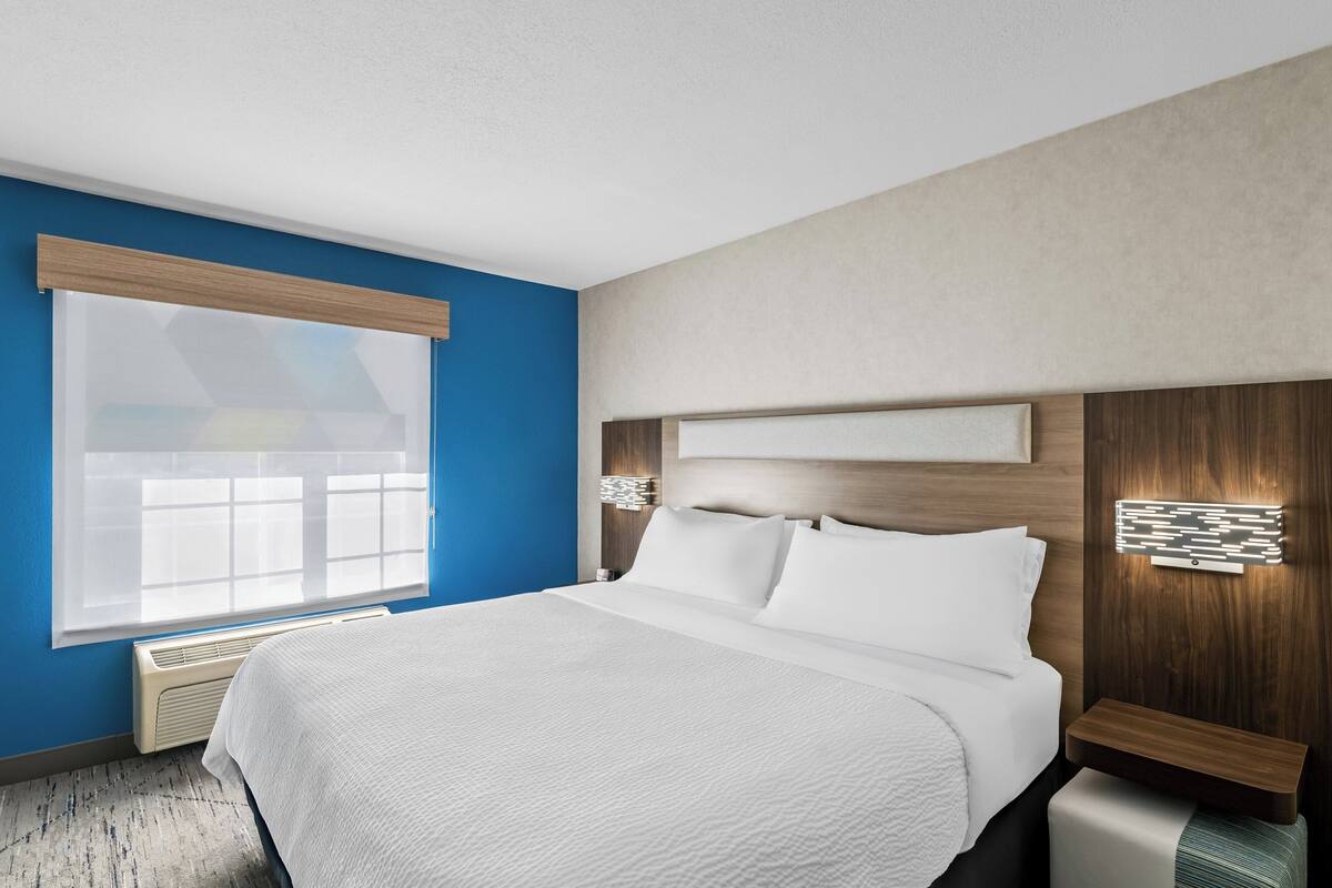 Une chambre d’hôtel moderne avec un grand lit, une fenêtre avec des stores, un luminaire fixé au mur et une tête de lit en bois.