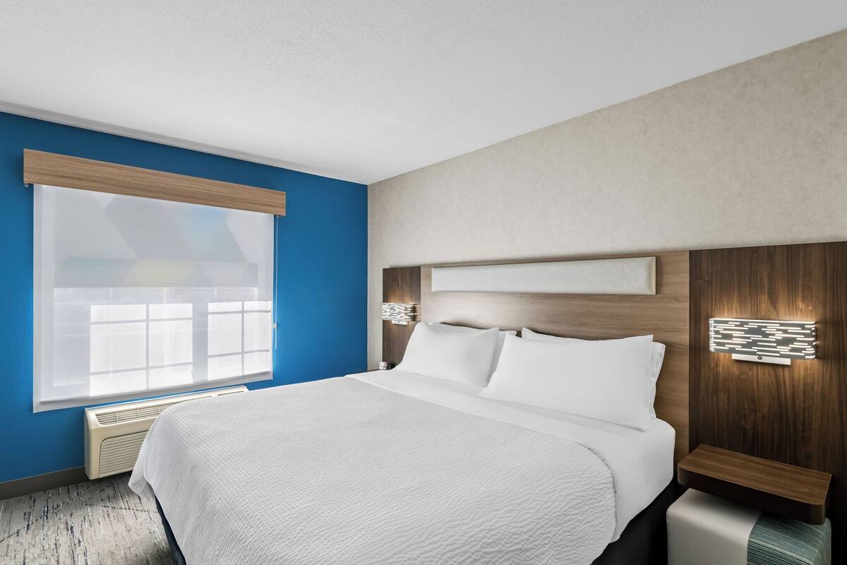 Une chambre d’hôtel avec un grand lit, une fenêtre avec des stores, un luminaire fixé au mur et une tête de lit en bois.