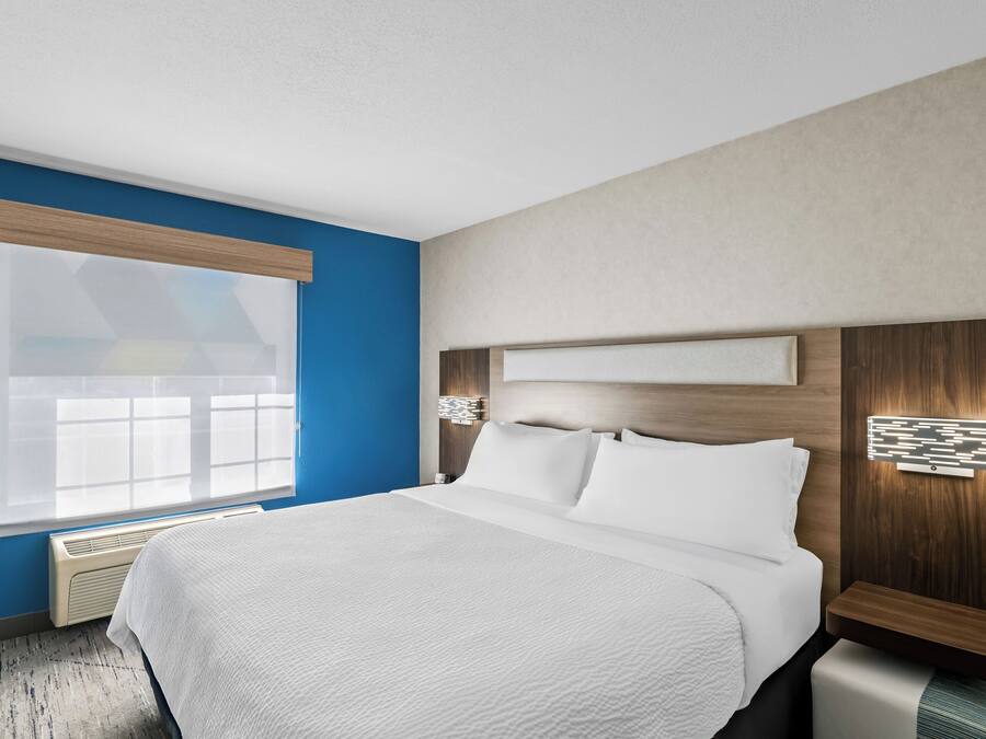 Quarto de hotel com uma cama grande, janela com persianas, luminária fixada na parede e cabeceira de madeira.