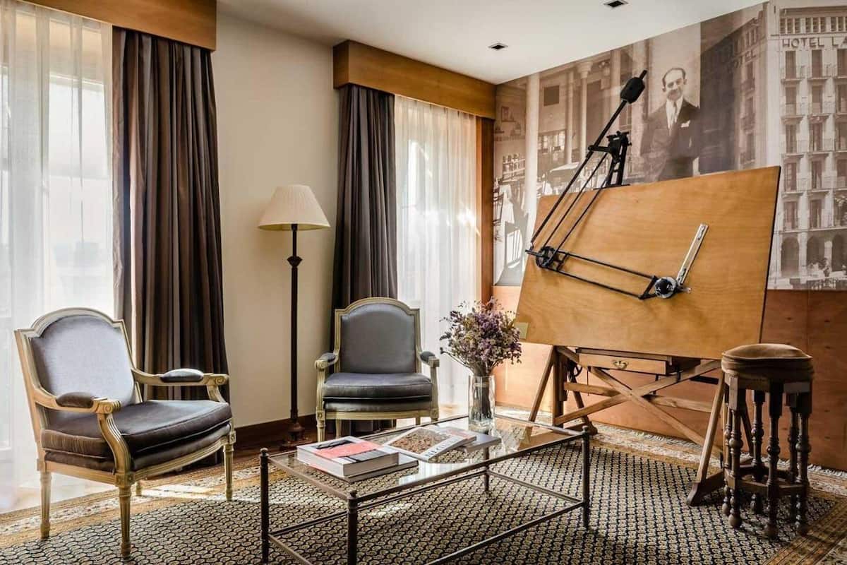 Habitación de hotel con diseño clásico, que incluye un ventanal con cortinas, una lámpara de pie, un caballete de madera con una pintura y una alfombra estampada.