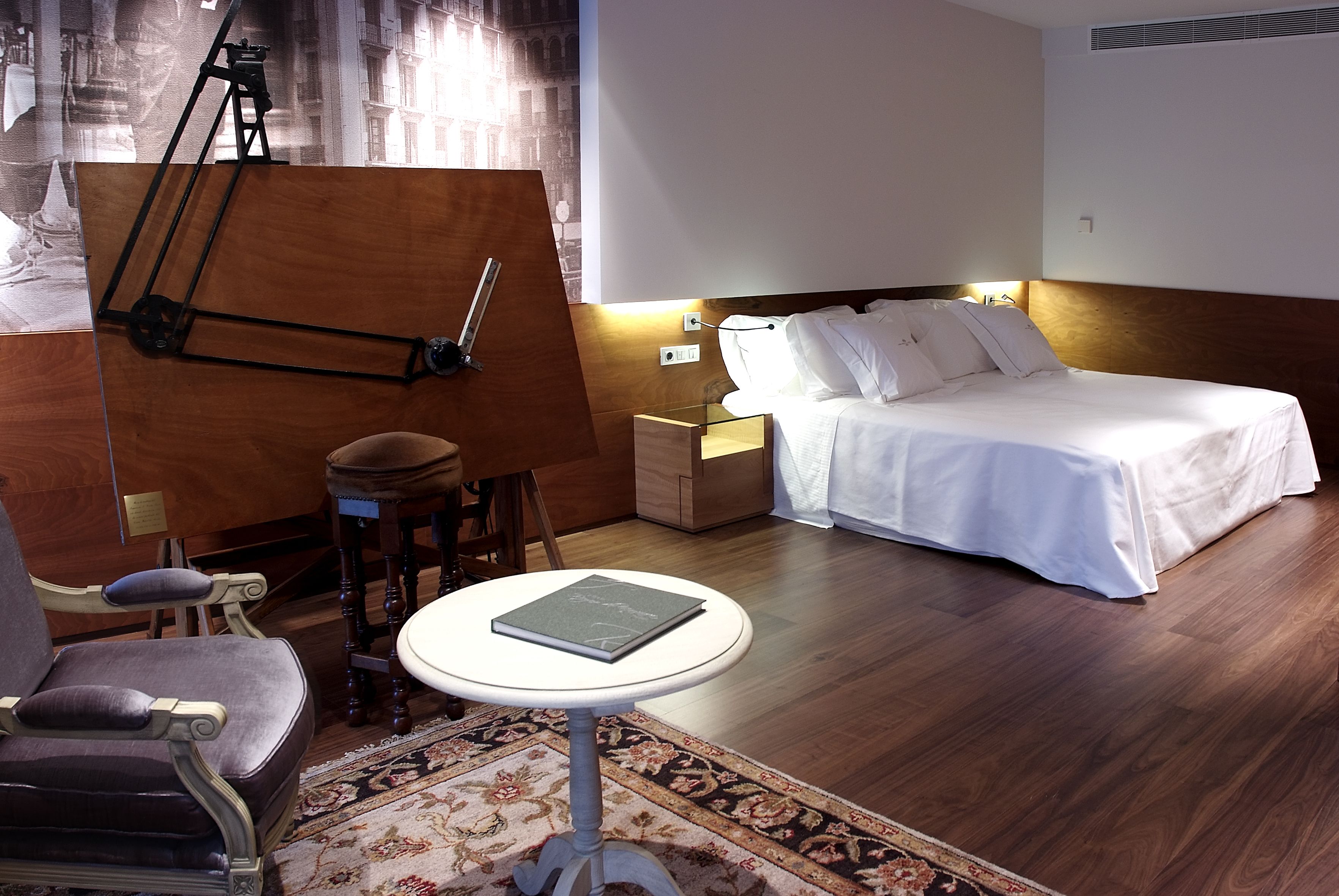 Habitació d'hotel amb un llit, una escriptori amb una cadira, una taula petita i una làmpada penjada a la paret.