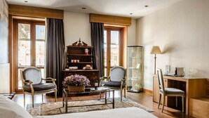 Habitación de hotel con una cama grande, dos sillones, una estantería de madera, un aparador de vidrio y un escritorio con lámpara.