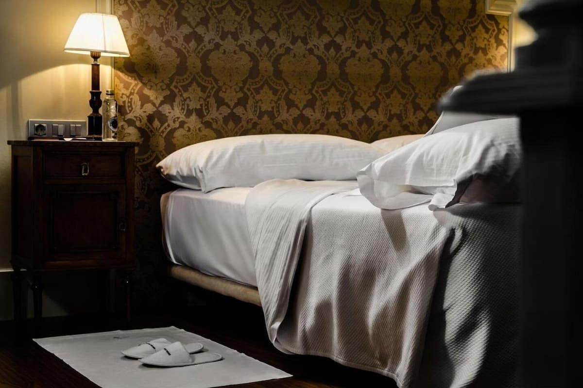 Una cama bien hecha con sábanas blancas, una lámpara de noche y una mesita de noche con una botella y un interruptor.