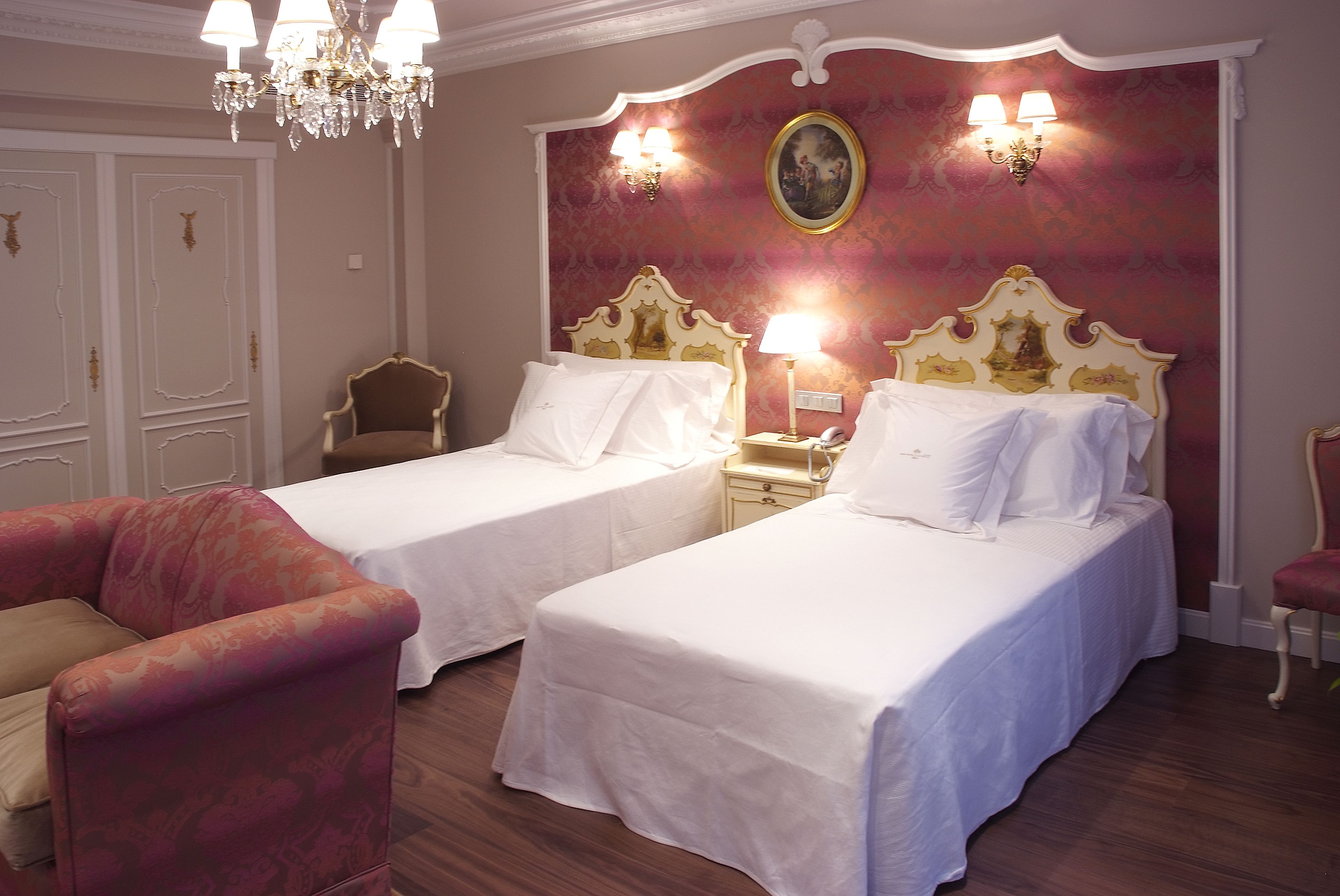 A hotel room with two beds, a chandelier, a red patterned wall, and a painting.