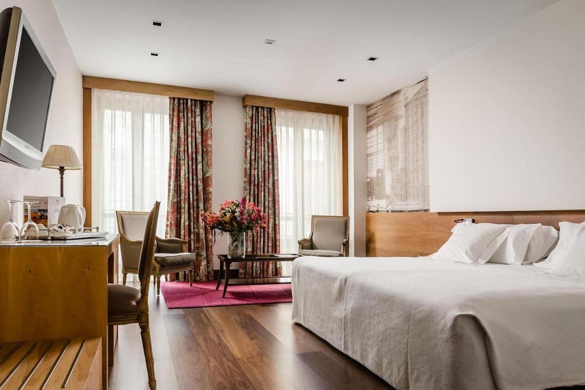 Habitación de hotel con cama, escritorio con lámpara, silla, pequeña mesa con un florero y un ventanal con cortinas.