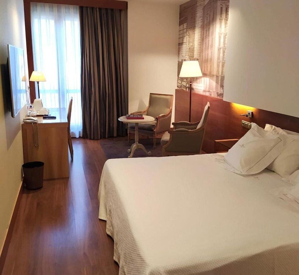 A hotel room with a large bed, a desk with a lamp, a chair, and a small table.