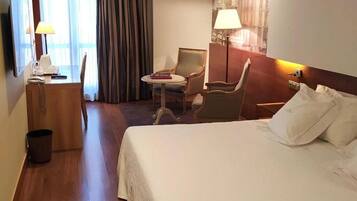 A hotel room with a large bed, a desk with a lamp, a chair, and a small table.