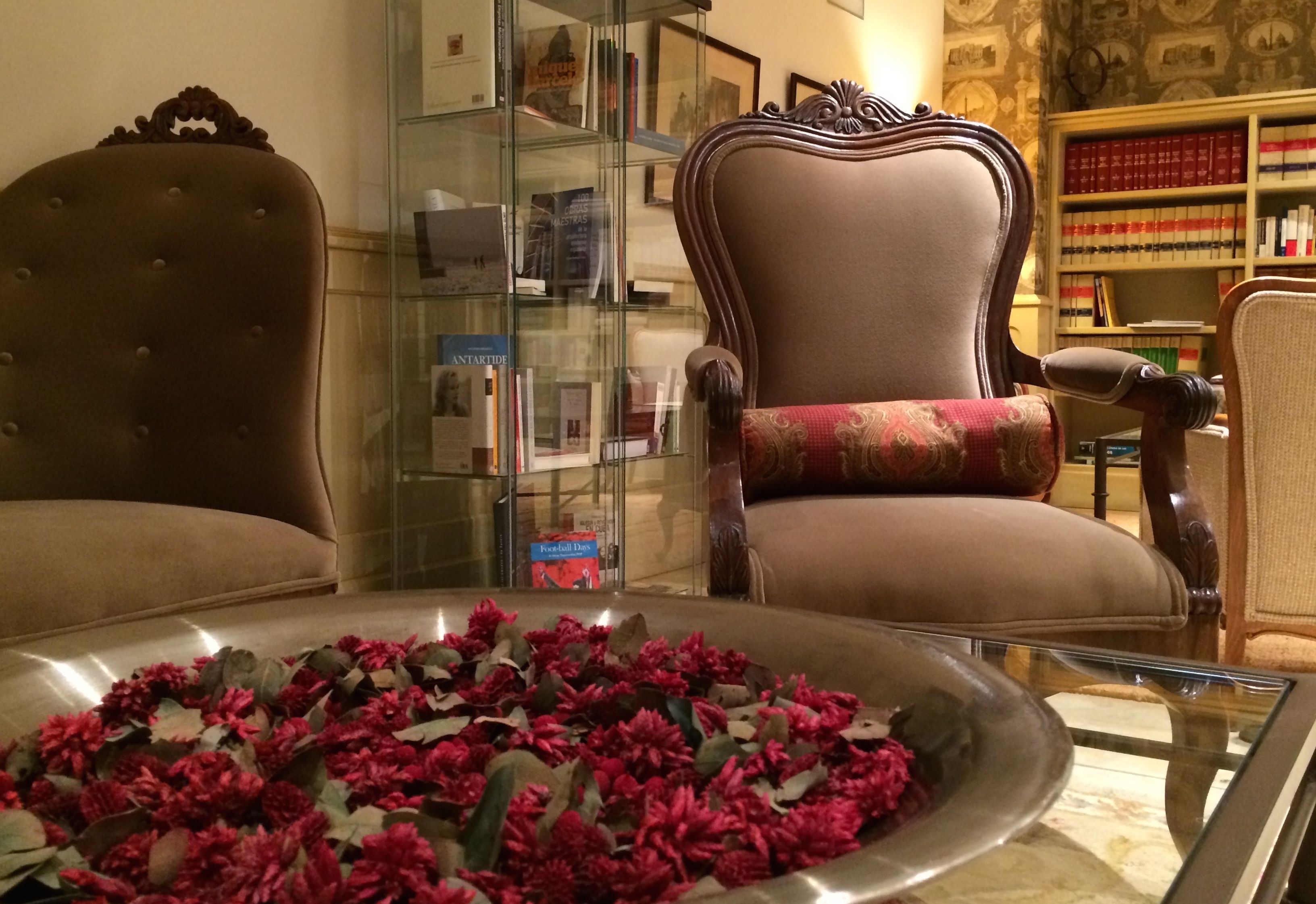 Una habitación con dos sillones tapizados, una vitrina de cristal con libros y una bandeja de flores rojas.