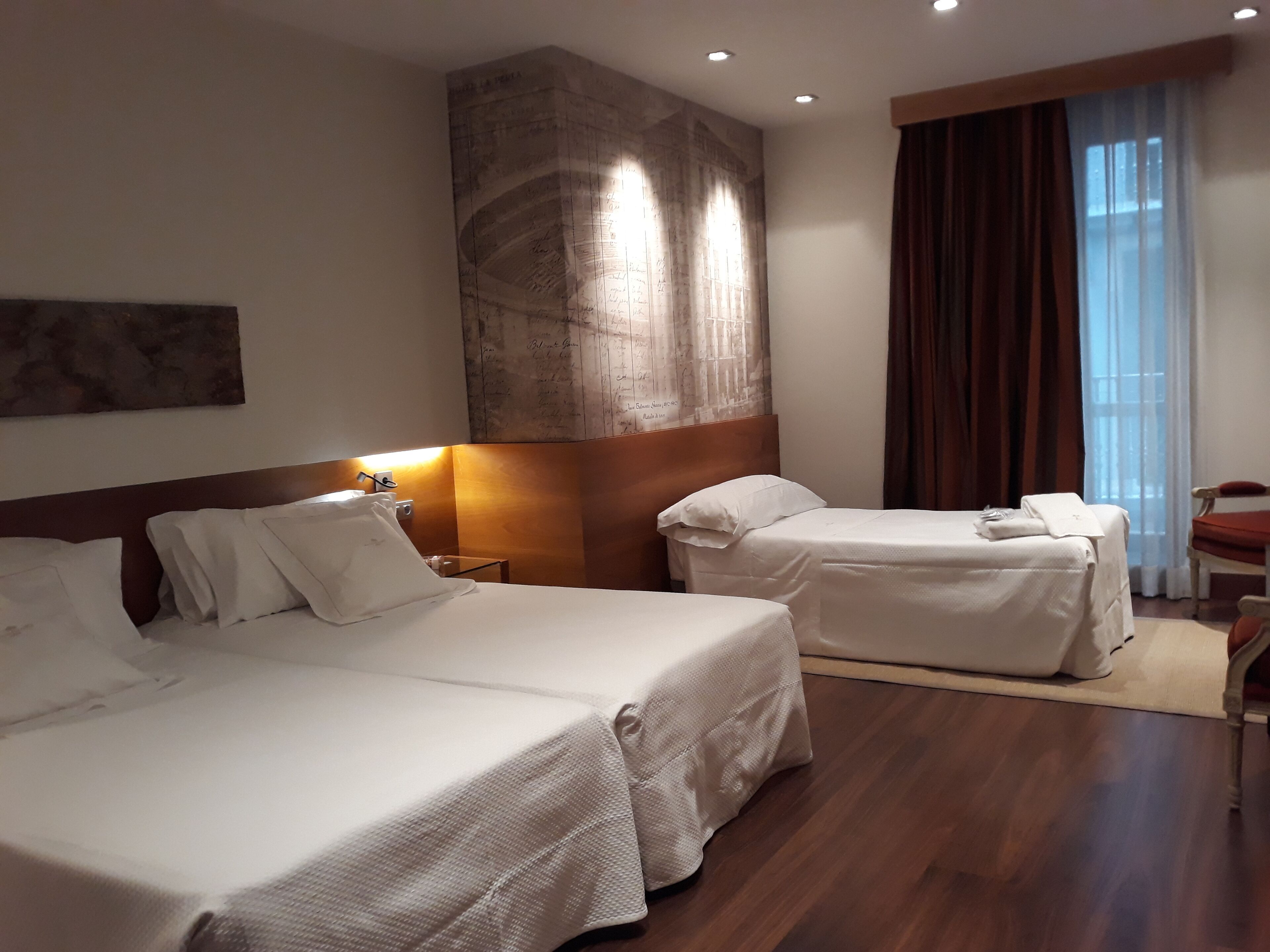 Habitación de hotel con dos camas, un mural grande en la pared, una mesita de noche y una ventana con cortinas.