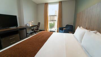 Habitación de hotel con una cama grande, un escritorio, un televisor de pantalla plana, una silla y vistas a edificios por la ventana.