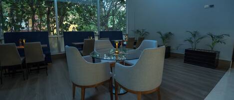 A modern dining area with a glass table surrounded by wicker chairs, large windows with a view of trees, and a wooden floor.