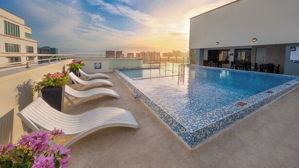 A rooftop pool with lounge chairs and a bar area, overlooking a cityscape at sunset.