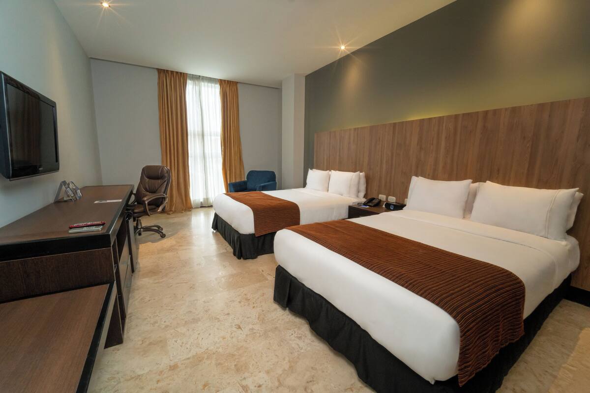 Ein Hotelzimmer mit zwei Betten, einem Schreibtisch, einem Sessel, einem Fernseher und einem Fenster mit Vorhängen.