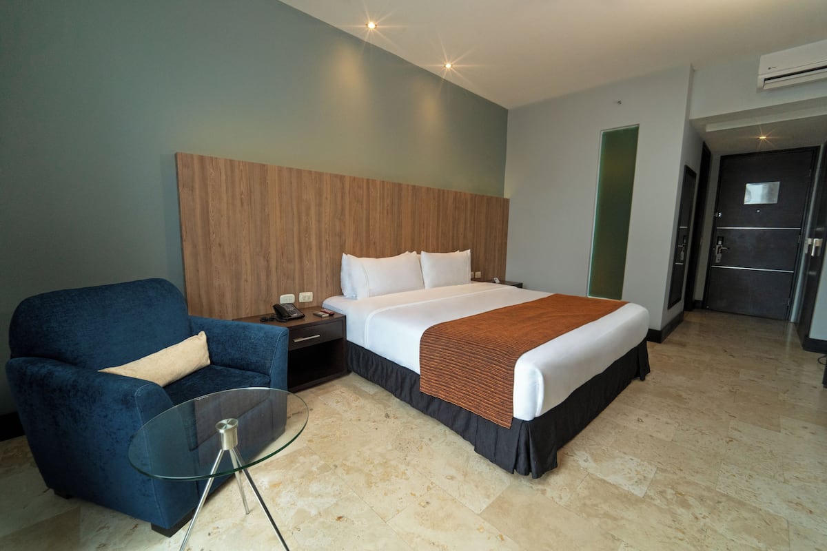 Ein Hotelzimmer mit einem Bett, einem blauen Sessel, einem Glastisch und einem Holzkopfteil.