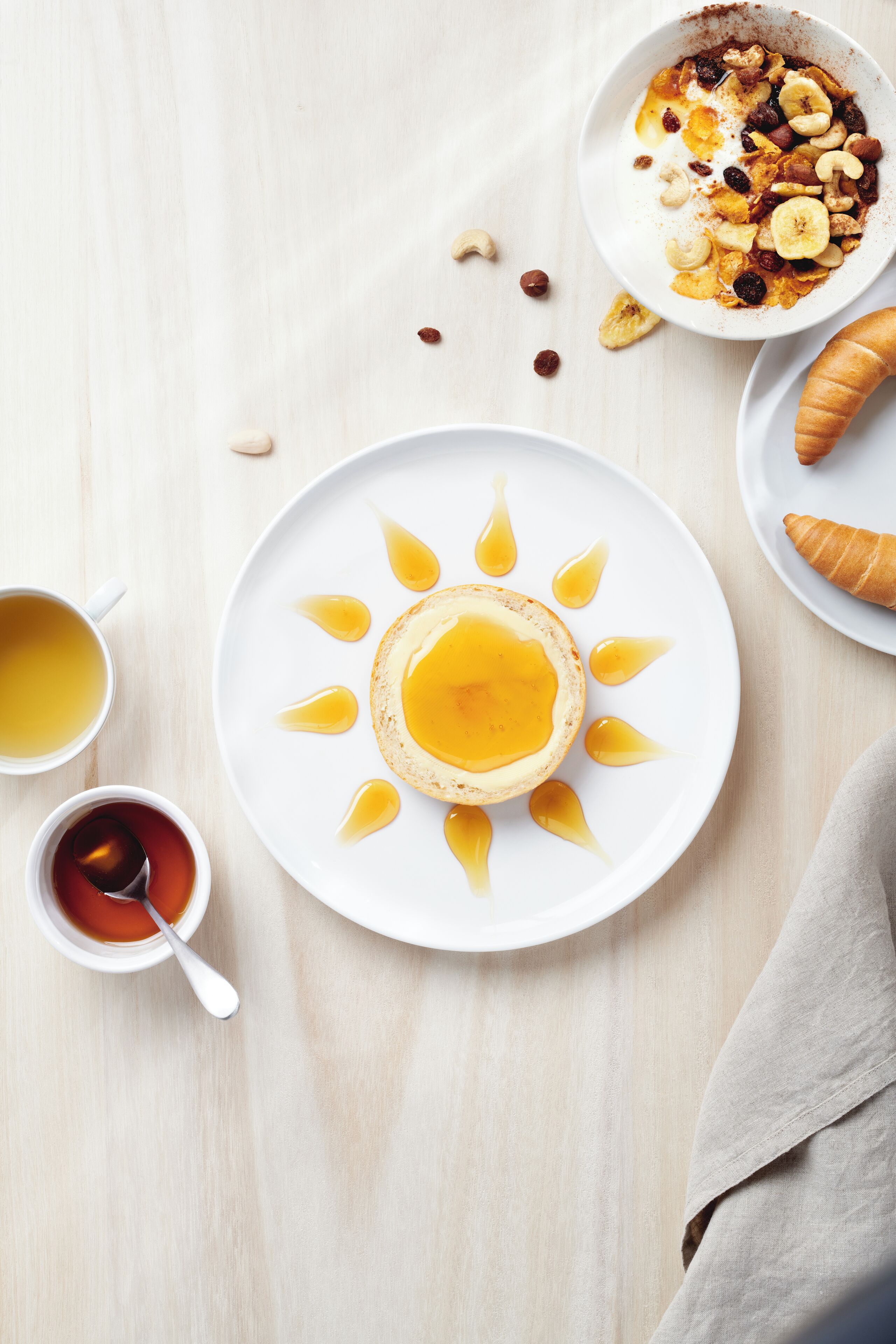 A breakfast setting with a plate of pancakes topped with syrup, a croissant, and a bowl of cereal with nuts and raisins. There are also two cups of tea and a napkin.