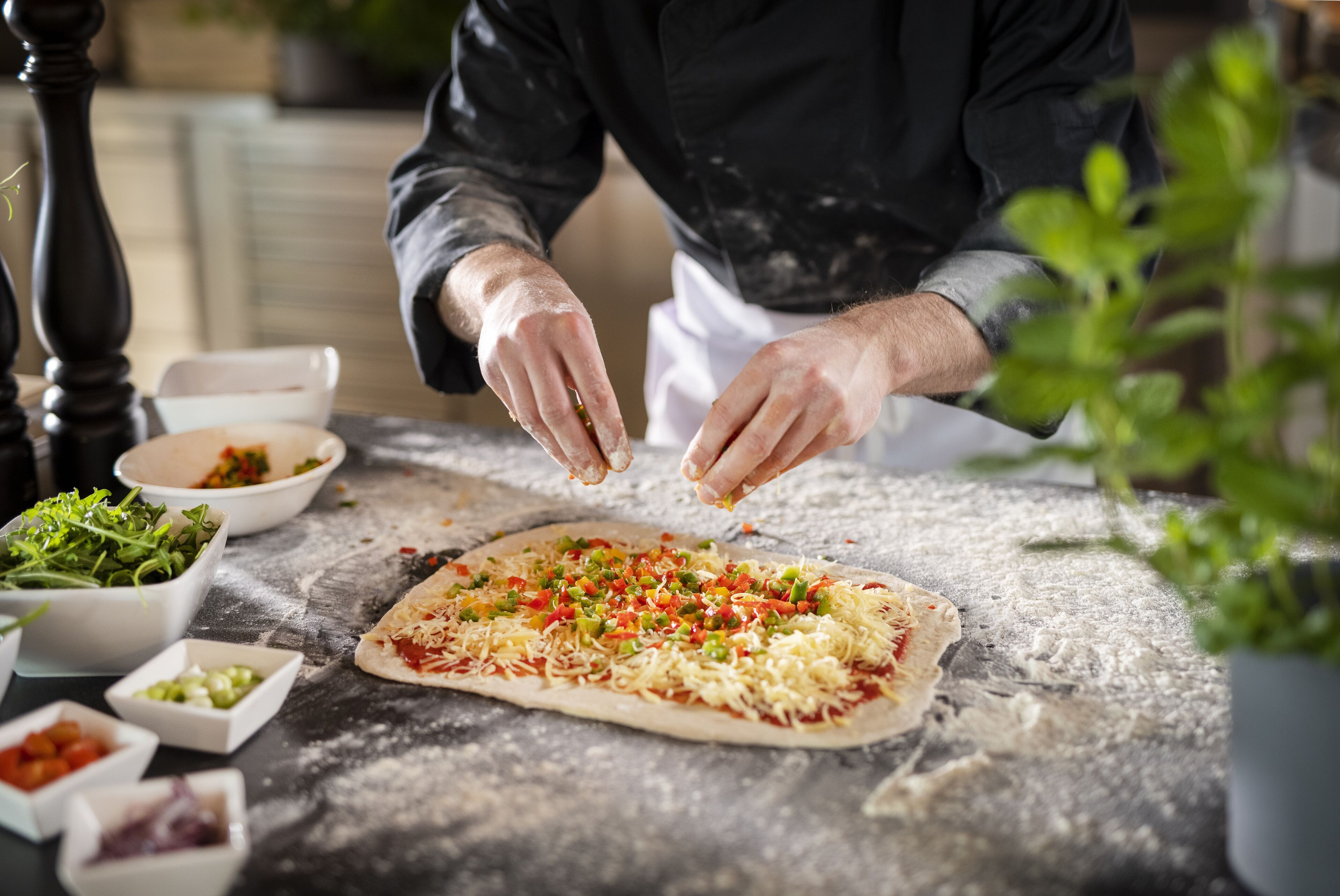 Eine Person bereitet Pizza an einem Tisch mit verschiedenen Zutaten und Schüsseln zu.