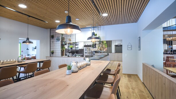 A modern dining area with a long wooden table, leather chairs, pendant lighting, and a kitchen in the background. - JUFA Hotel Wipptal (Steinach am Brenner)