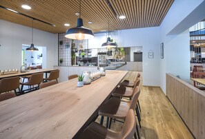 A modern dining area with a long wooden table, leather chairs, pendant lighting, and a kitchen in the background. - JUFA Hotel Wipptal (Steinach am Brenner)
