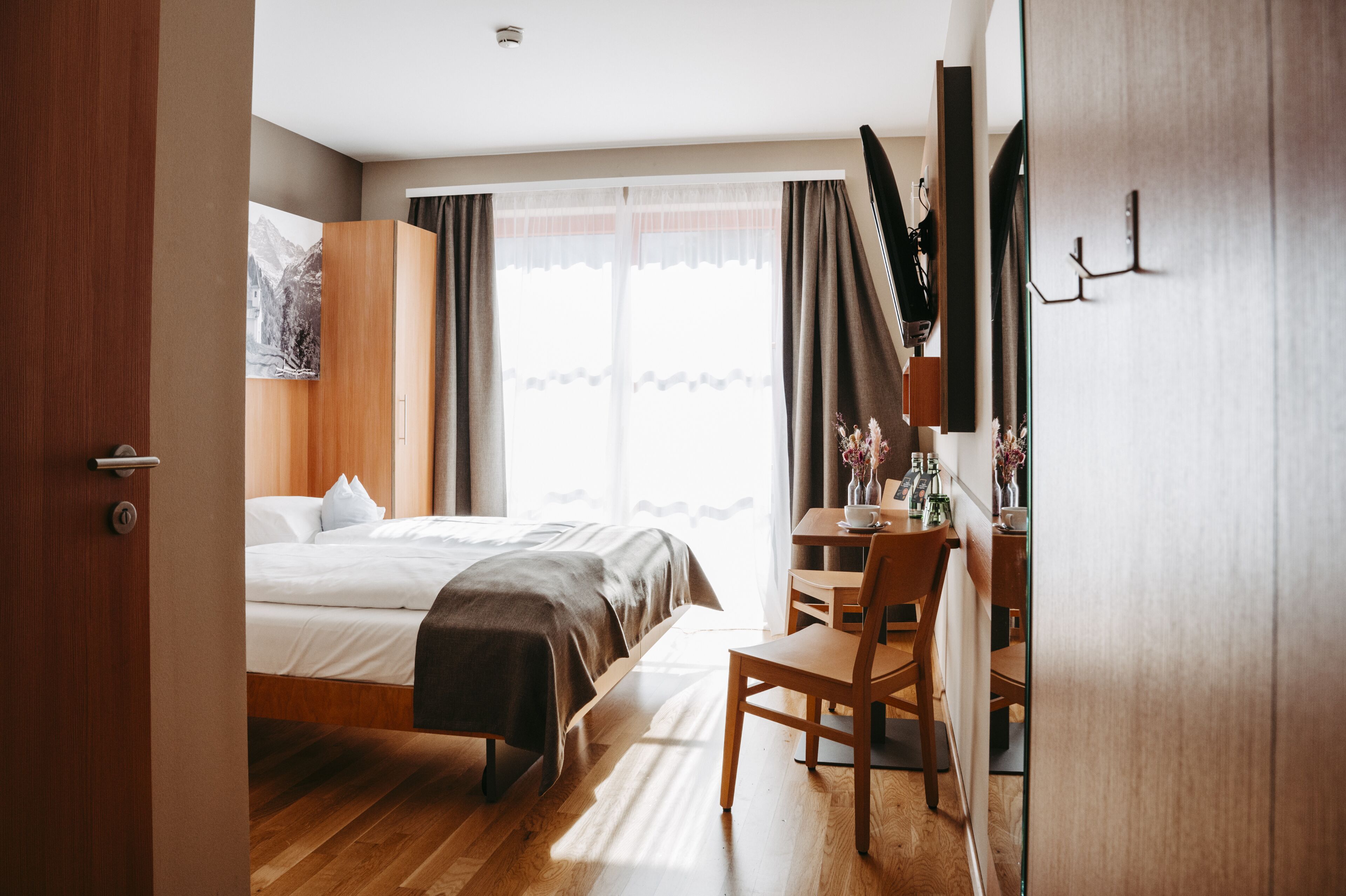 Habitación de hotel con una cama, un escritorio y una silla. Hay una ventana con cortinas y un cuadro en la pared.