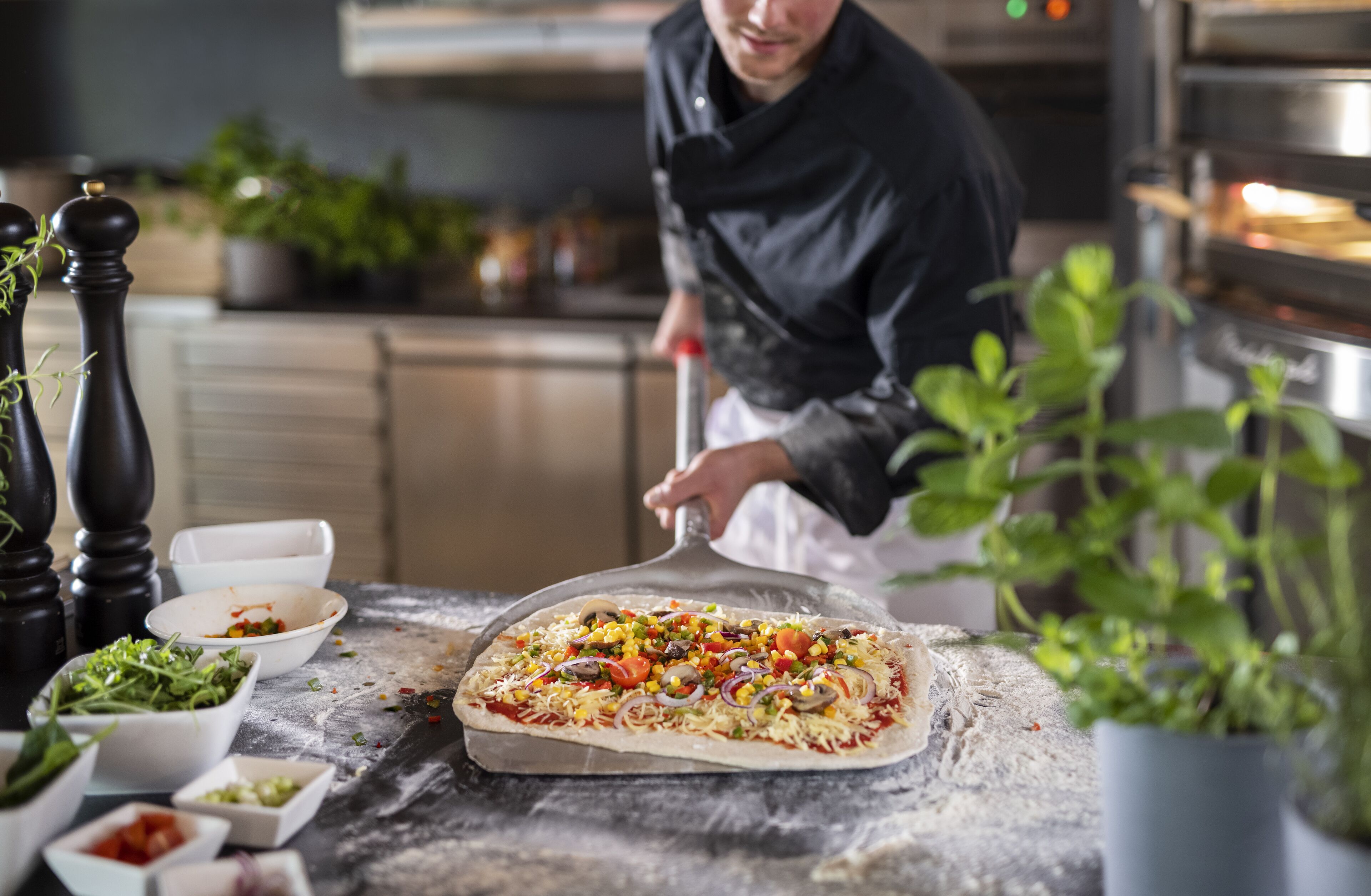En kock som förbereder en pizza på en träskärbräda i ett kök med olika ingredienser och köksredskap.