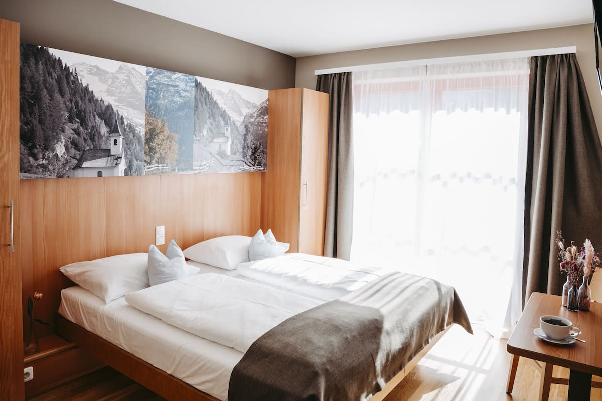 A hotel room with a bed, a wooden wardrobe, a table with a cup, and a window with curtains.