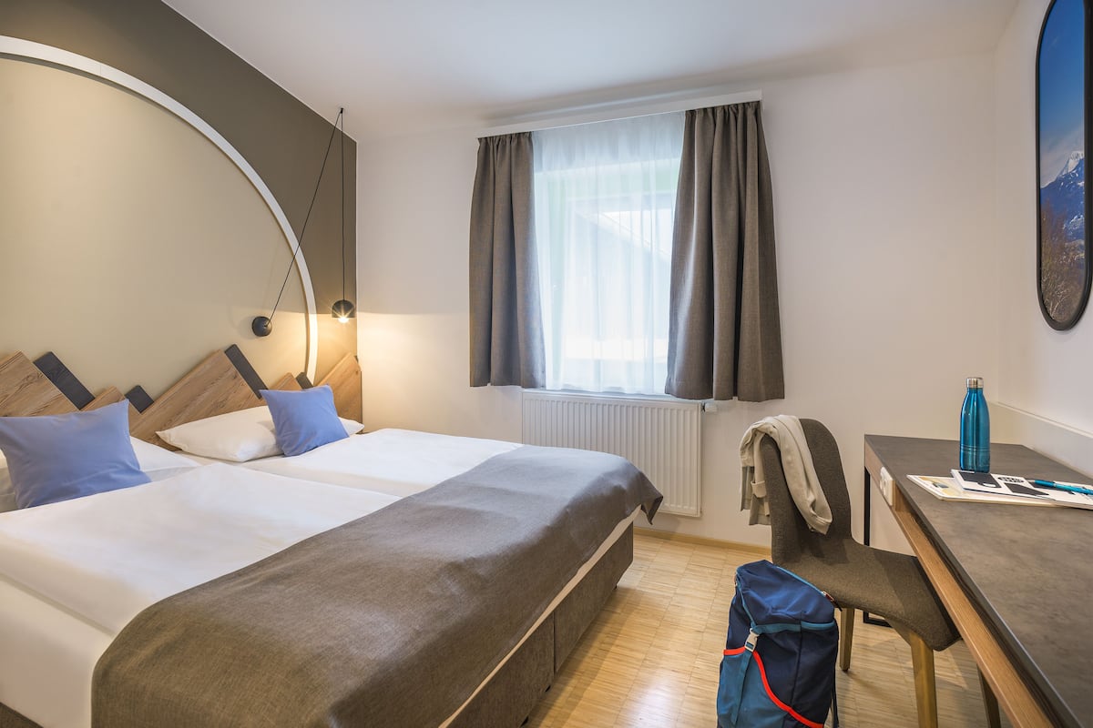 A hotel room with a large bed, a desk, and a chair. There is a window with curtains, a lamp, and a backpack on the floor.