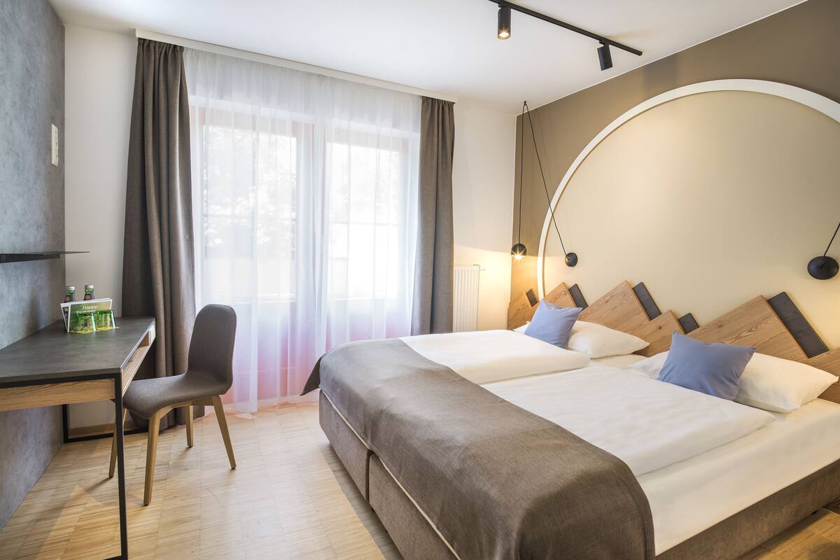 A hotel room with two beds, a desk, a chair, and a large window with curtains.