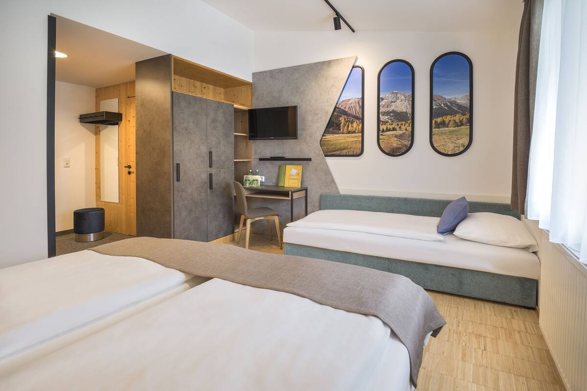 A hotel room with two beds, a desk, a chair, a TV, and a large window with a mountain view.
