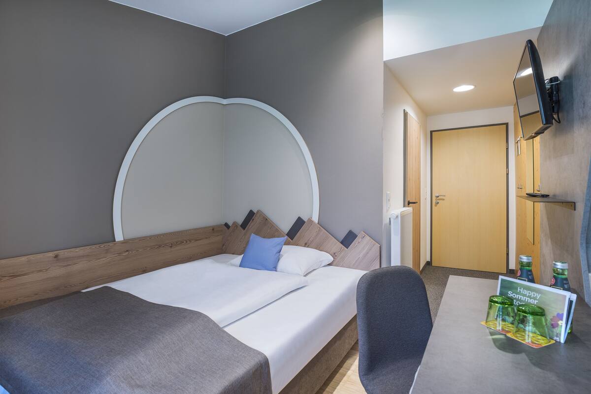 A hotel room with a bed, a desk, a chair, a TV mounted on the wall, and a small table with a 'Happy Summer' sign.