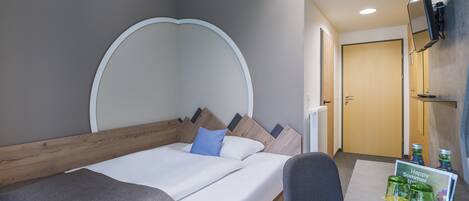 Habitación de hotel con una cama, un escritorio, una silla, un televisor montado en la pared y una mesita con un cartel que dice 'Feliz Verano'.