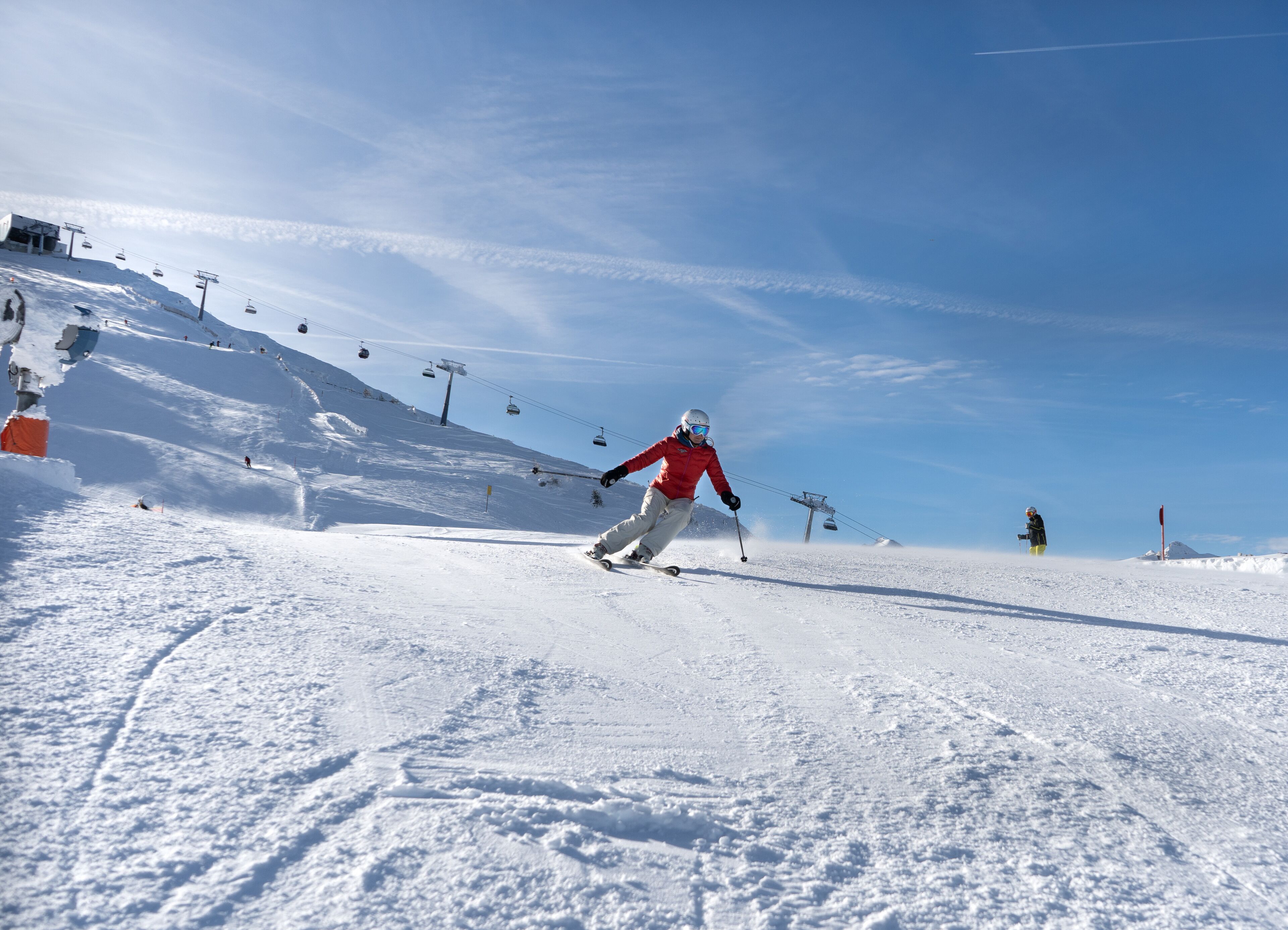 Eine Skipiste mit einer Person, die abfährt, Seilbahnen im Hintergrund und einem klaren Himmel.