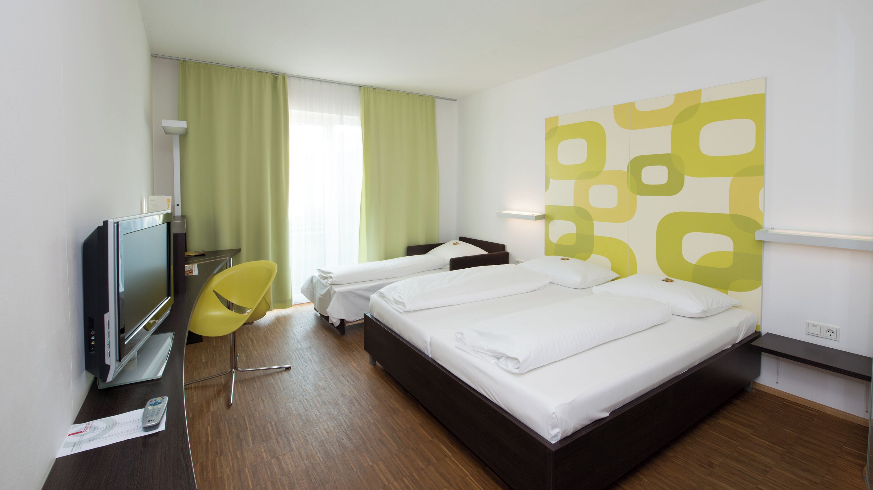 A hotel room with two single beds, a TV, a desk with a chair, and a large window with curtains.
