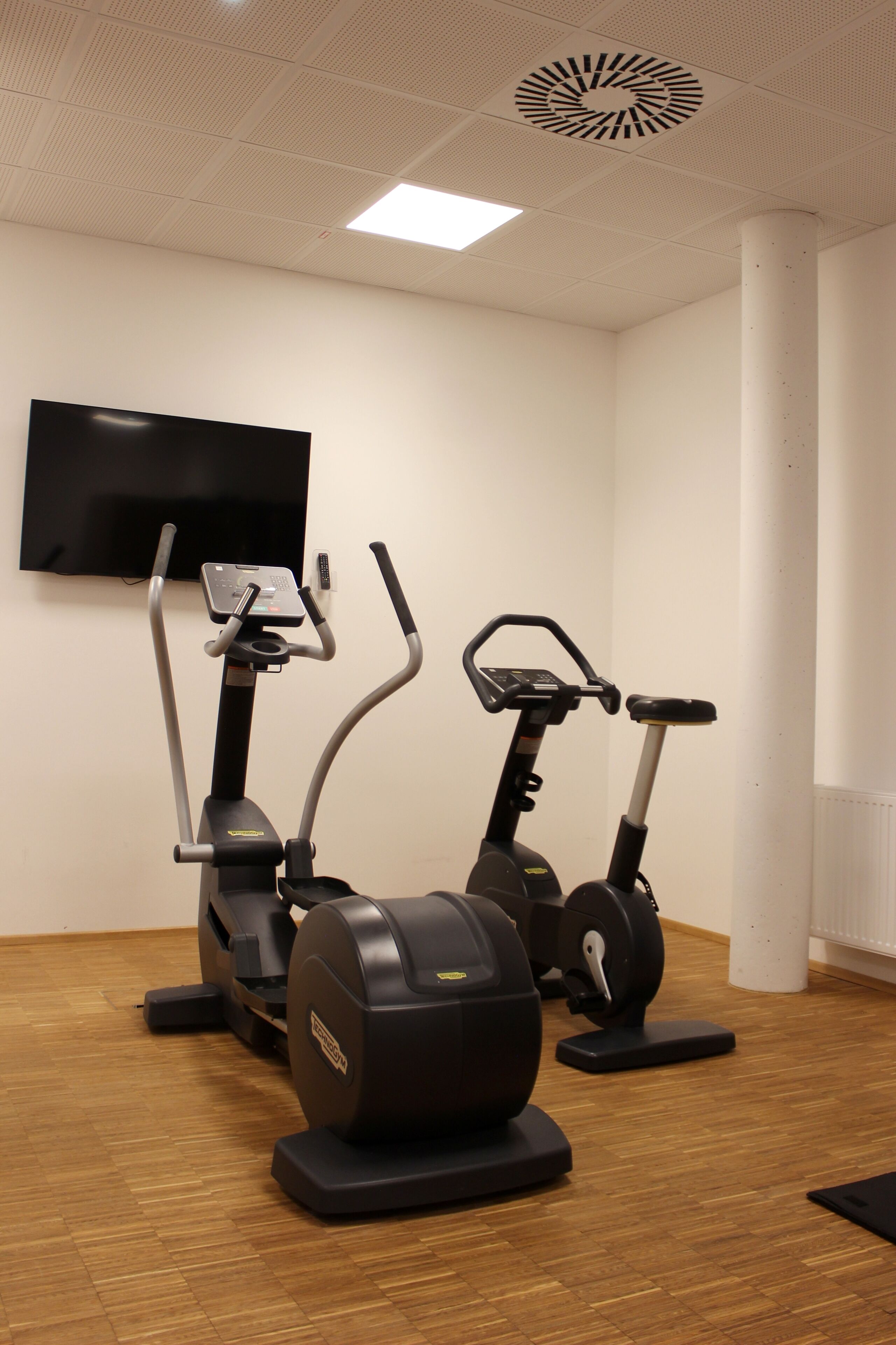 Una habitación con dos bicicletas estáticas, un televisor en la pared y un respiradero en el techo.
