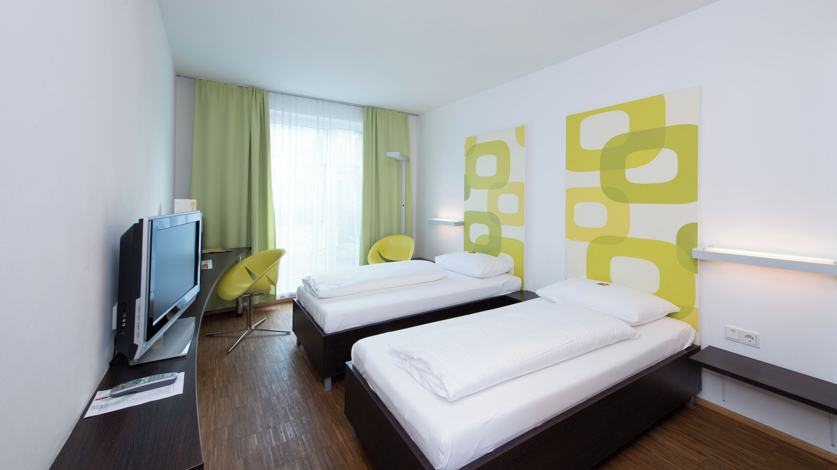 Une chambre d’hôtel avec deux lits, un bureau avec une télévision, une chaise et une fenêtre avec des rideaux.