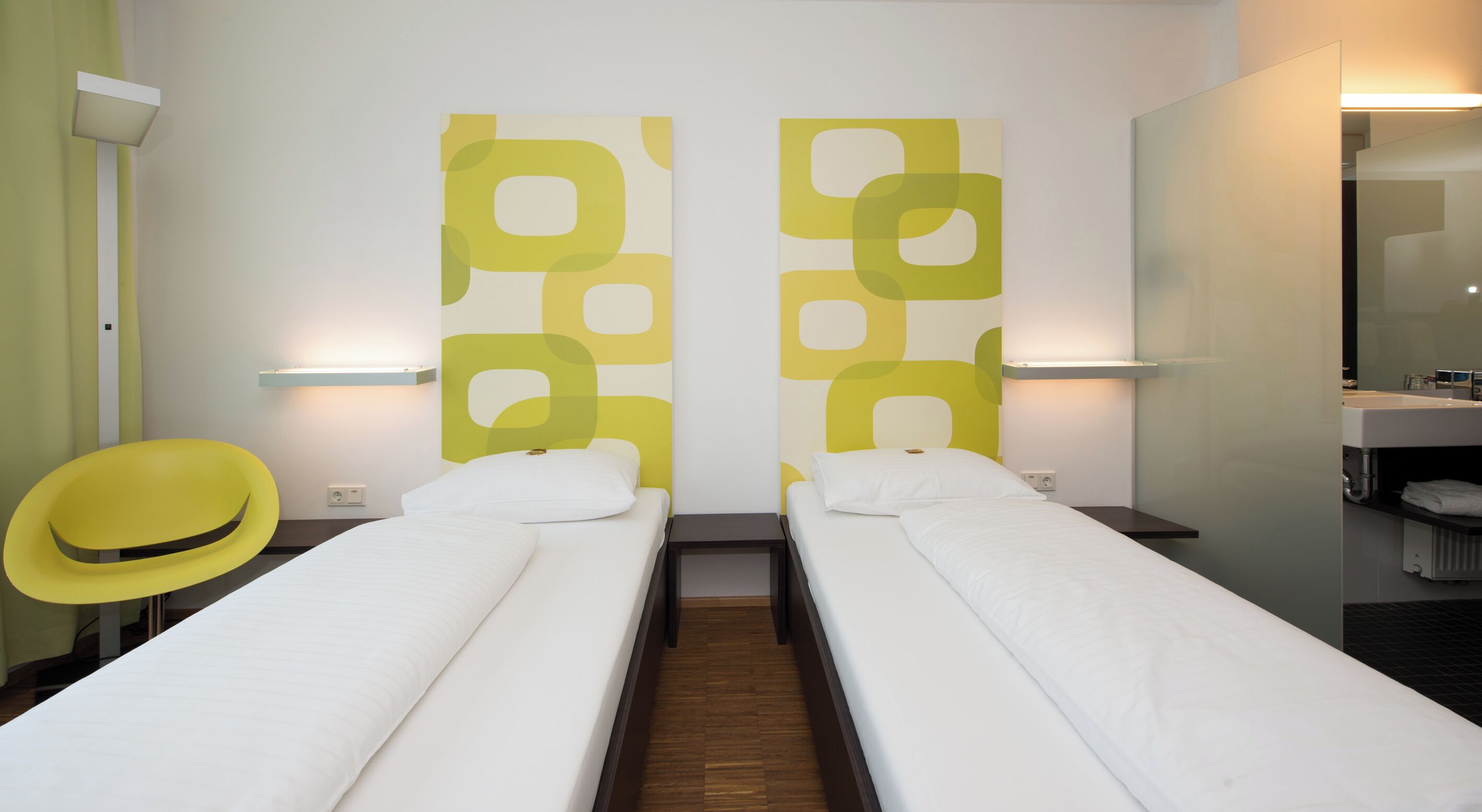 A hotel room with two single beds, a yellow chair, a large mirror, and a wall with abstract patterns.