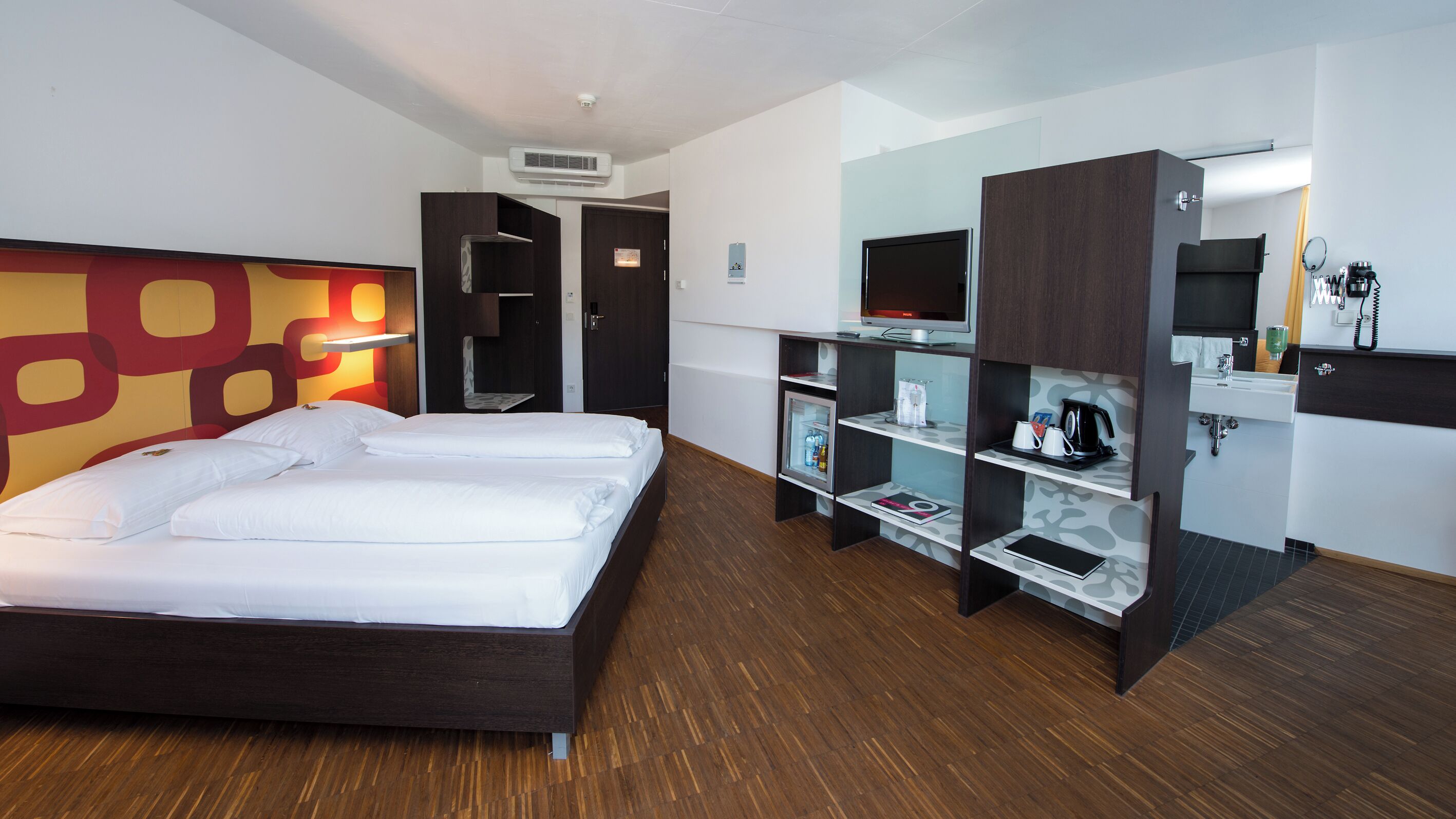 A hotel room with a large bed, a wardrobe with shelves, a mirror, a television, and a small refrigerator.