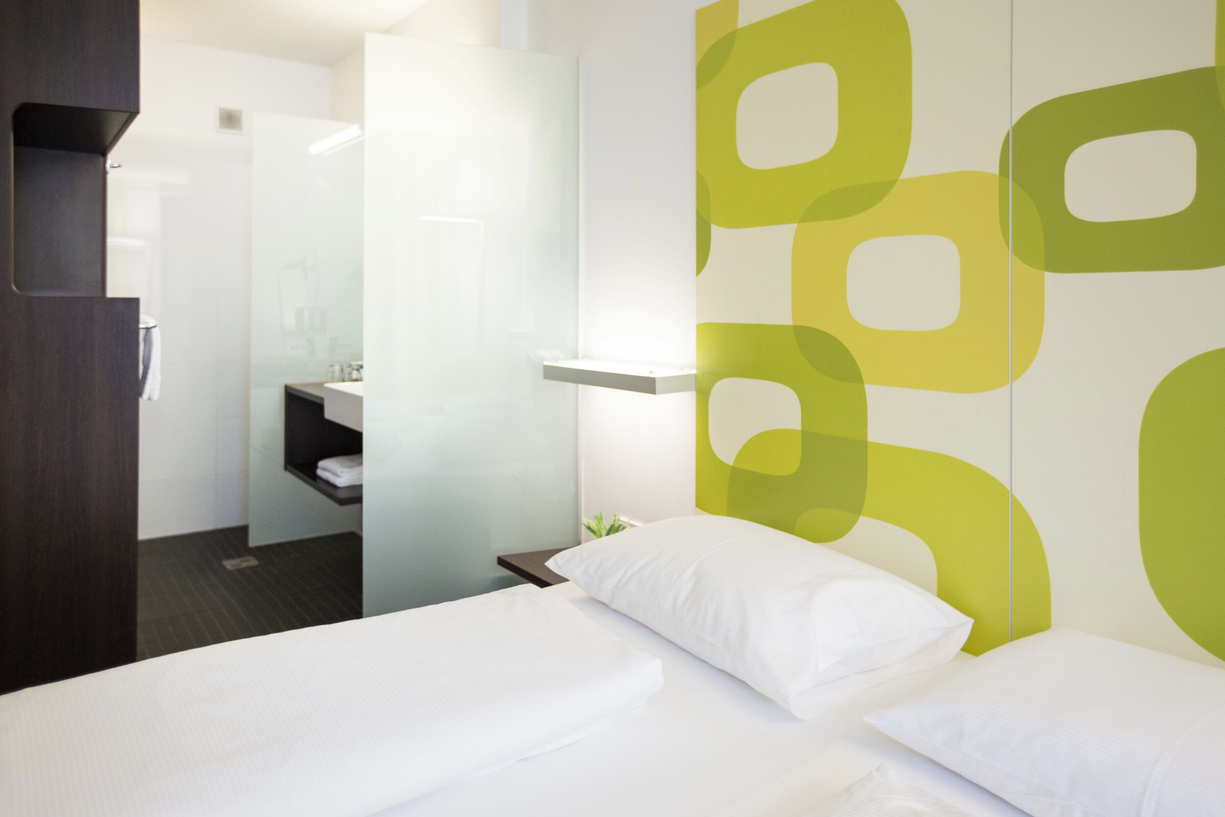 Una habitación de hotel moderna con una cama, una mesita de noche y un baño visible a través de un panel de vidrio.
