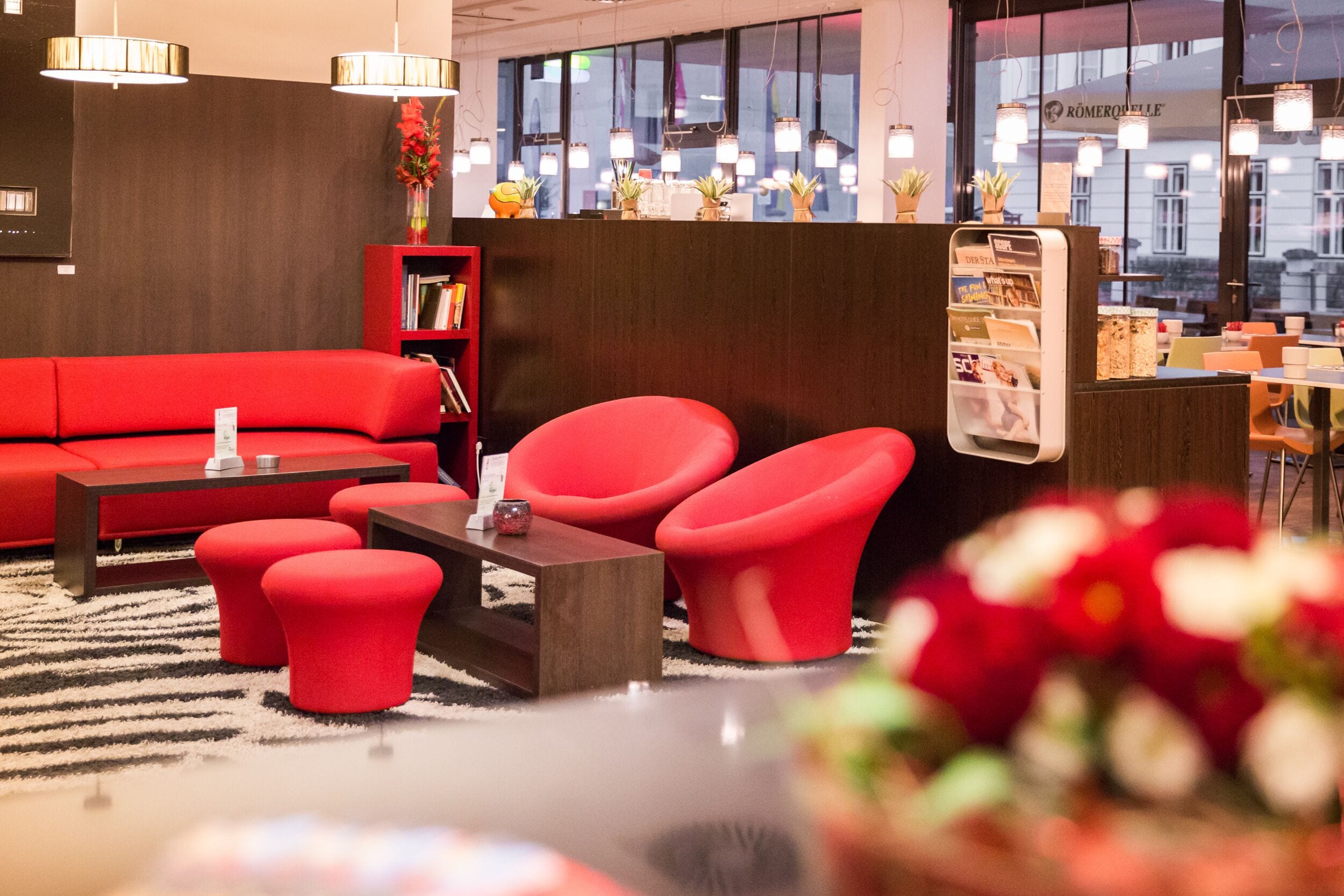 Un vestíbulo moderno con un sofá rojo, sillones a juego y una mesa de centro de madera. Se observa una estantería con libros y un jarrón con flores.