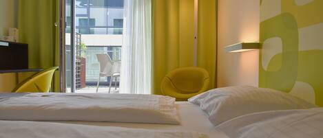 A hotel room with a bed, a yellow chair, a table with a lamp, and a view of the outside balcony.