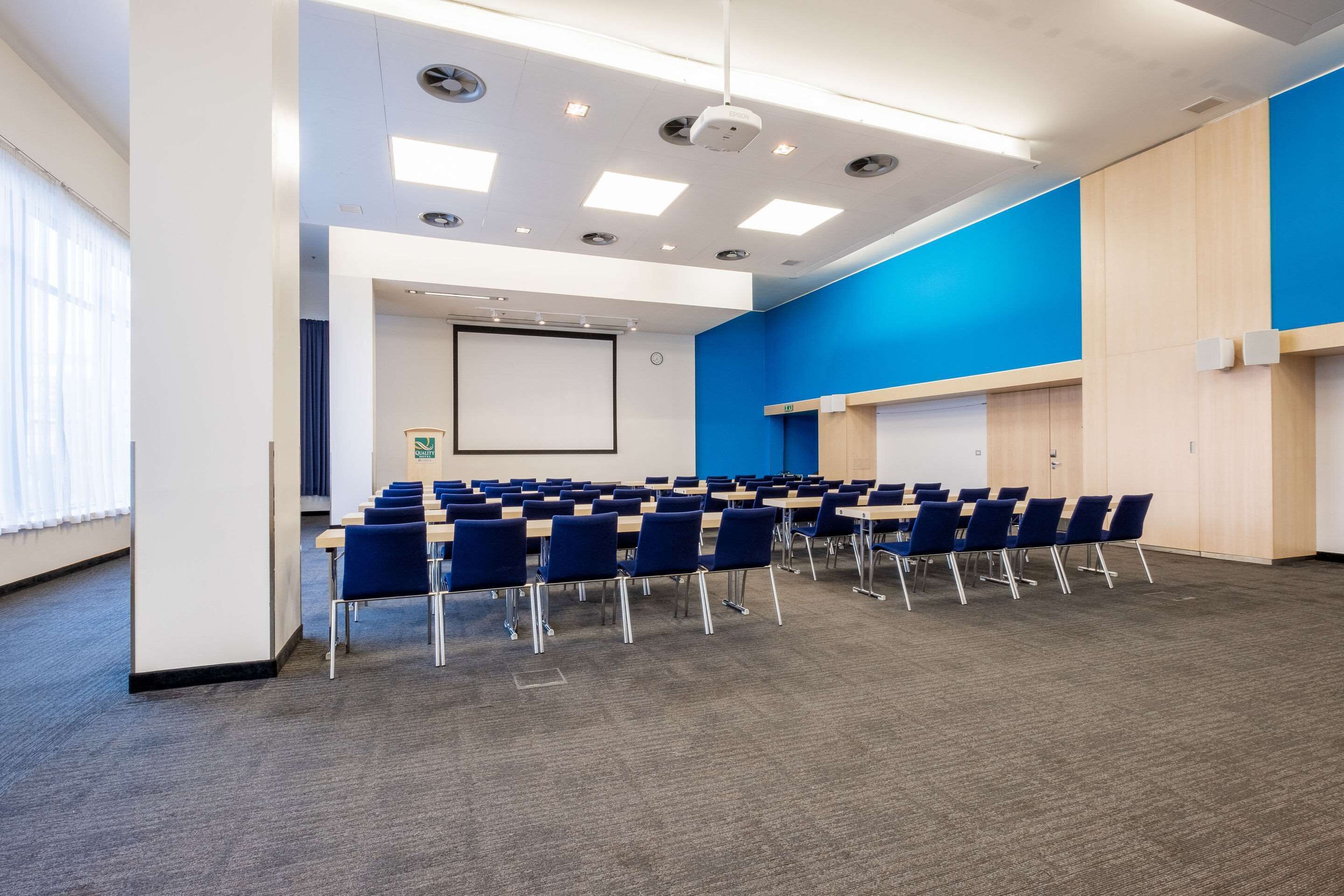 A conference room with rows of blue chairs, a projector screen, and a whiteboard.