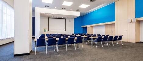 A conference room with rows of blue chairs, a projector screen, and a whiteboard.
