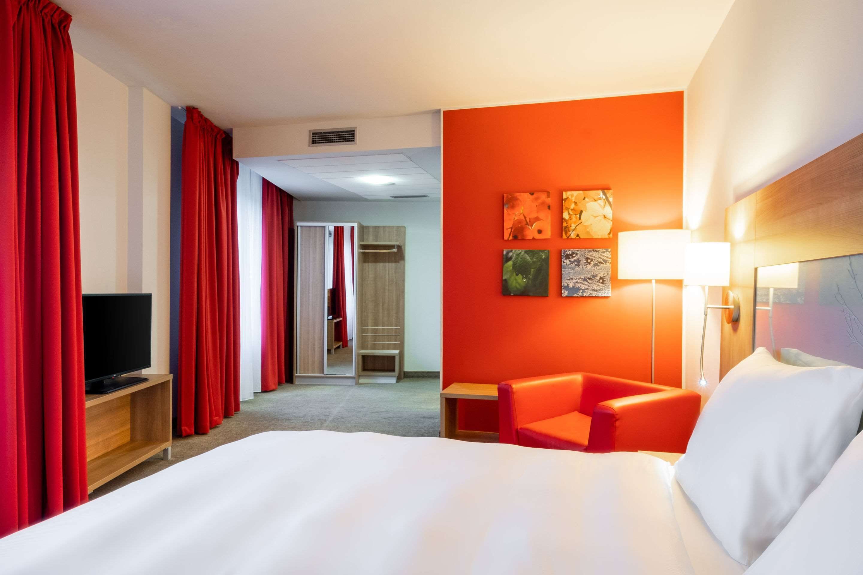 Habitación de hotel con una cama grande, cortinas rojas, un televisor sobre un atril de madera, un sillón rojo y una pared con tres cuadros enmarcados.